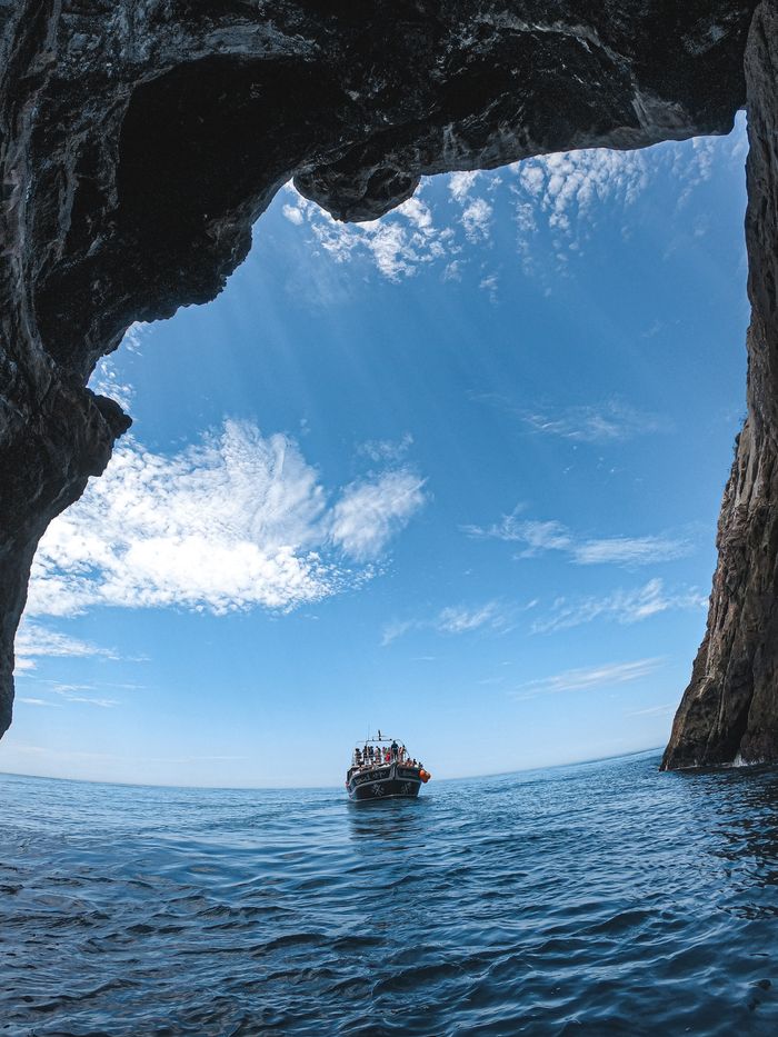 A boat navigates through crystal clear ocean waters towards a stunning cave opening against a bright blue sky.
